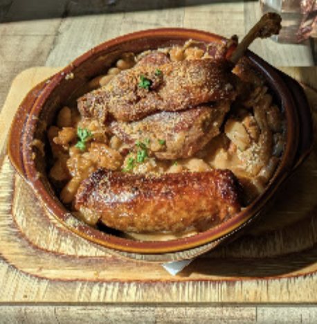 Confit et saucisse dorés dans une cassole en terre cuite, servie sur une planche.